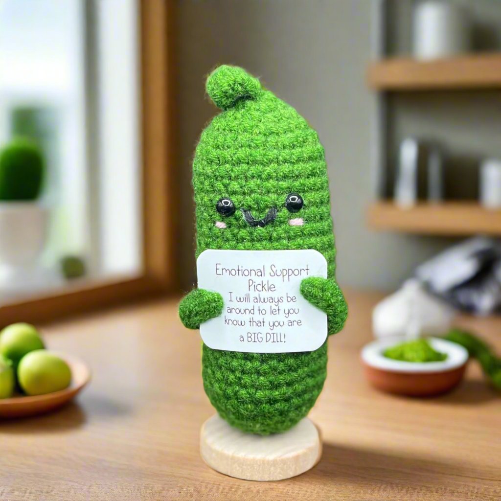 A crocheted, green pickle figurine with a smiling face, holding a sign that reads 'Emotional Support Pickle: I will always be around to let you know that you are a BIG DILL!' The figurine is placed on a wooden base.
